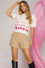 Woman wearing a white sweater with pink heart pattern and beige shorts against a pink and gray background