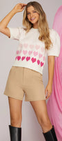 Woman wearing a white sweater with pink heart pattern and beige shorts against a pink and gray background