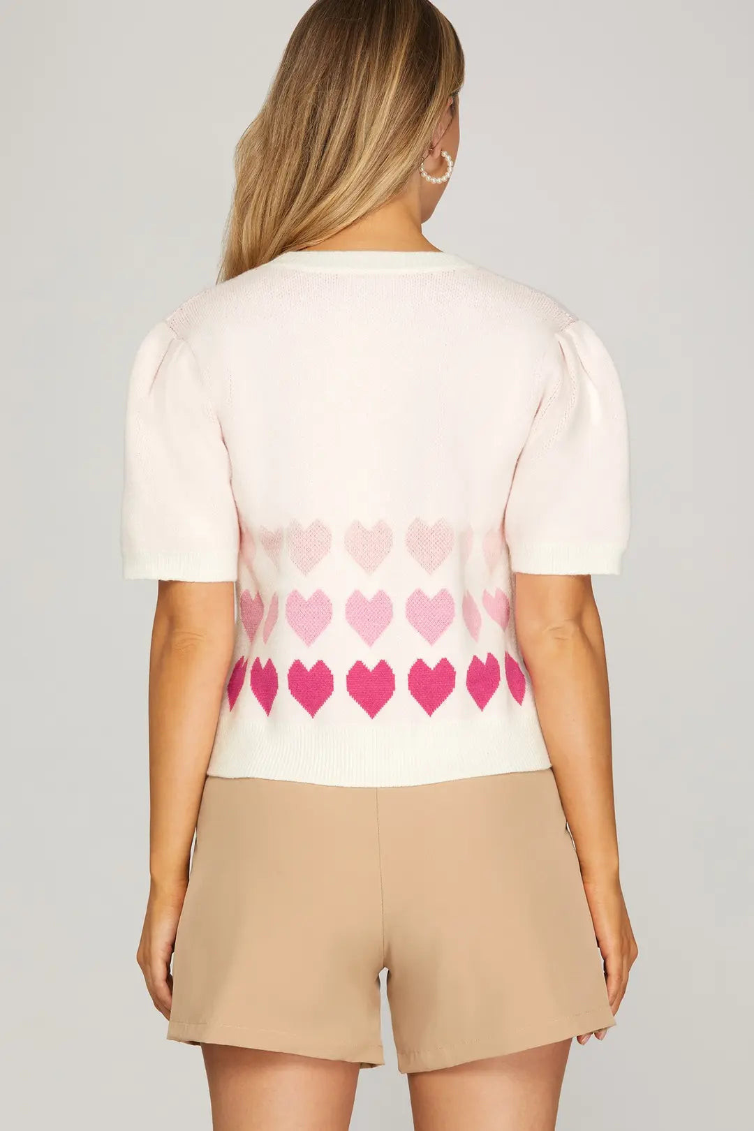 Woman wearing a white t-shirt with pink heart patterns and beige shorts on a gray background