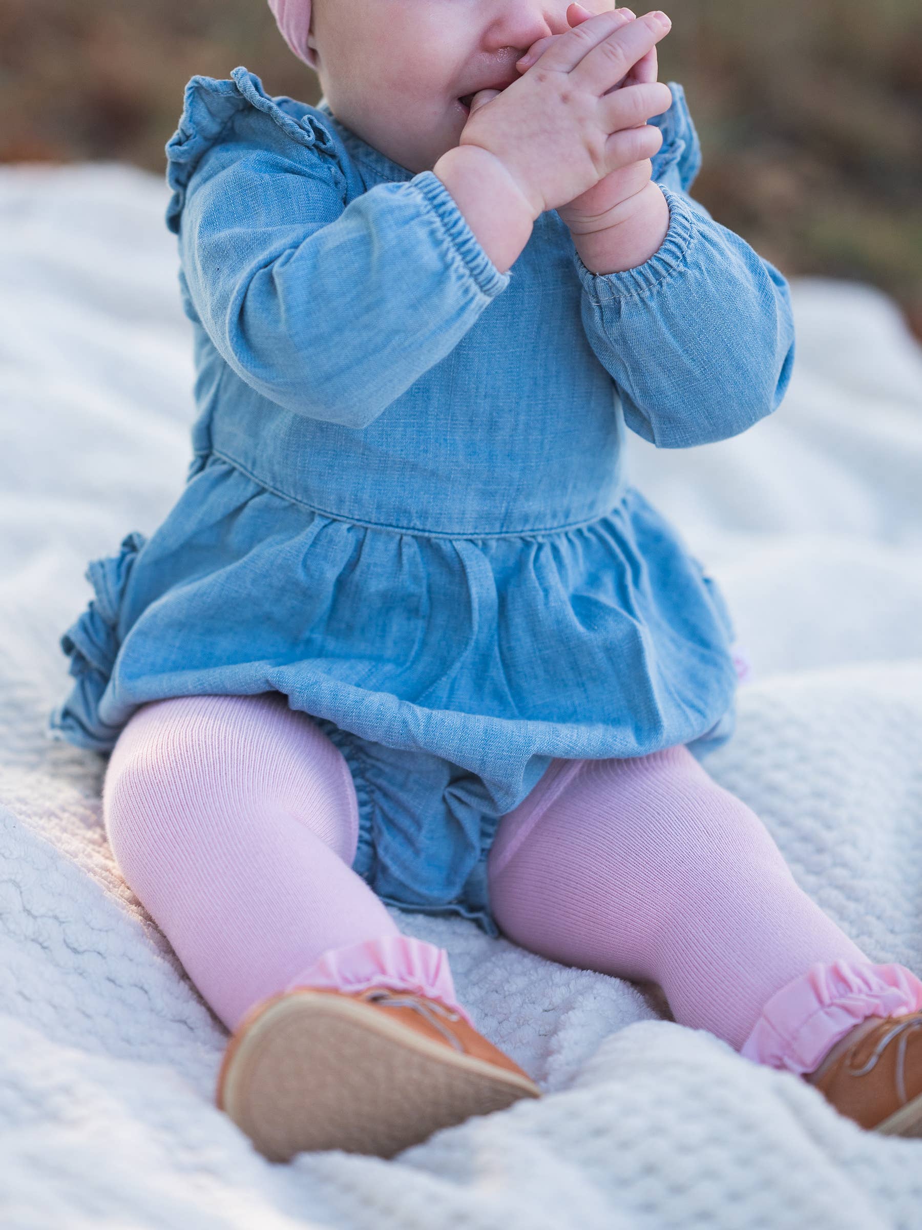 Baby in a blue dress and pink tights sitting on a blanket.