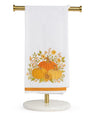 A white kitchen towel with an embroidered autumn harvest design featuring pumpkins, sunflowers, and leaves, hanging from a gold metal stand on a white background.