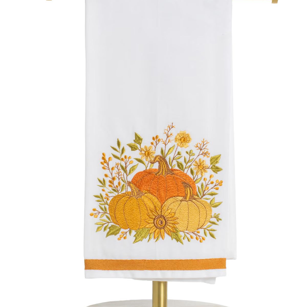 A white kitchen towel with an embroidered autumn harvest design featuring pumpkins, sunflowers, and leaves, hanging from a gold metal stand on a white background.