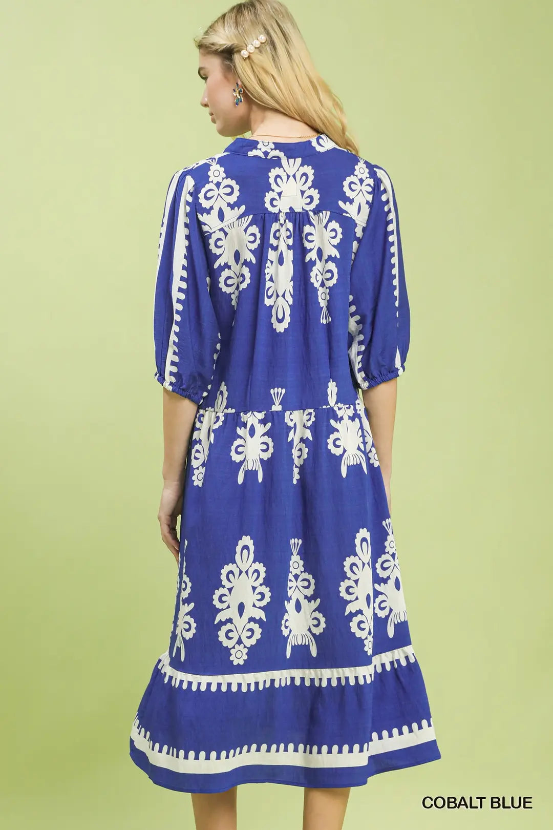 A woman shown from the back wearing a cobalt blue dress with white floral and ornamental patterns, photographed against a solid light green background.