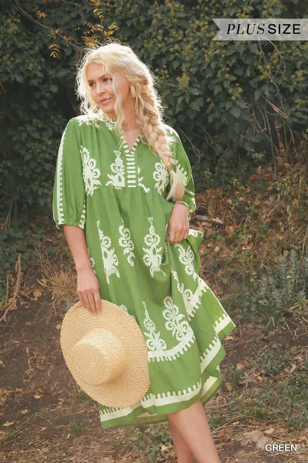 Woman wearing a green dress with white embroidery, holding a straw hat, standing outdoors.
