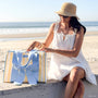 Woman in white dress and sun hat sitting on a beach with a striped bag.