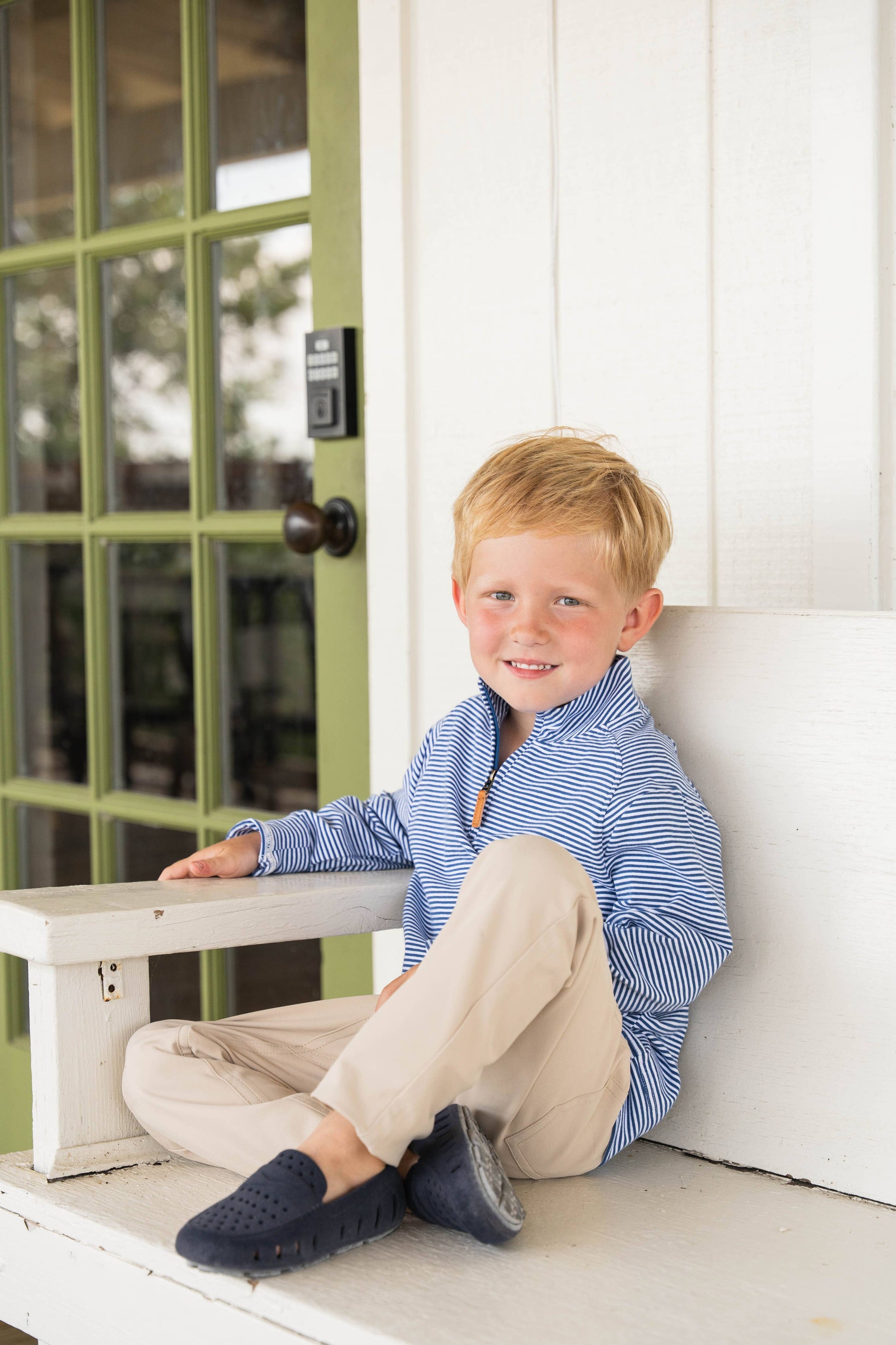 Young boy sitting on a step in front of a white door with green window frames.