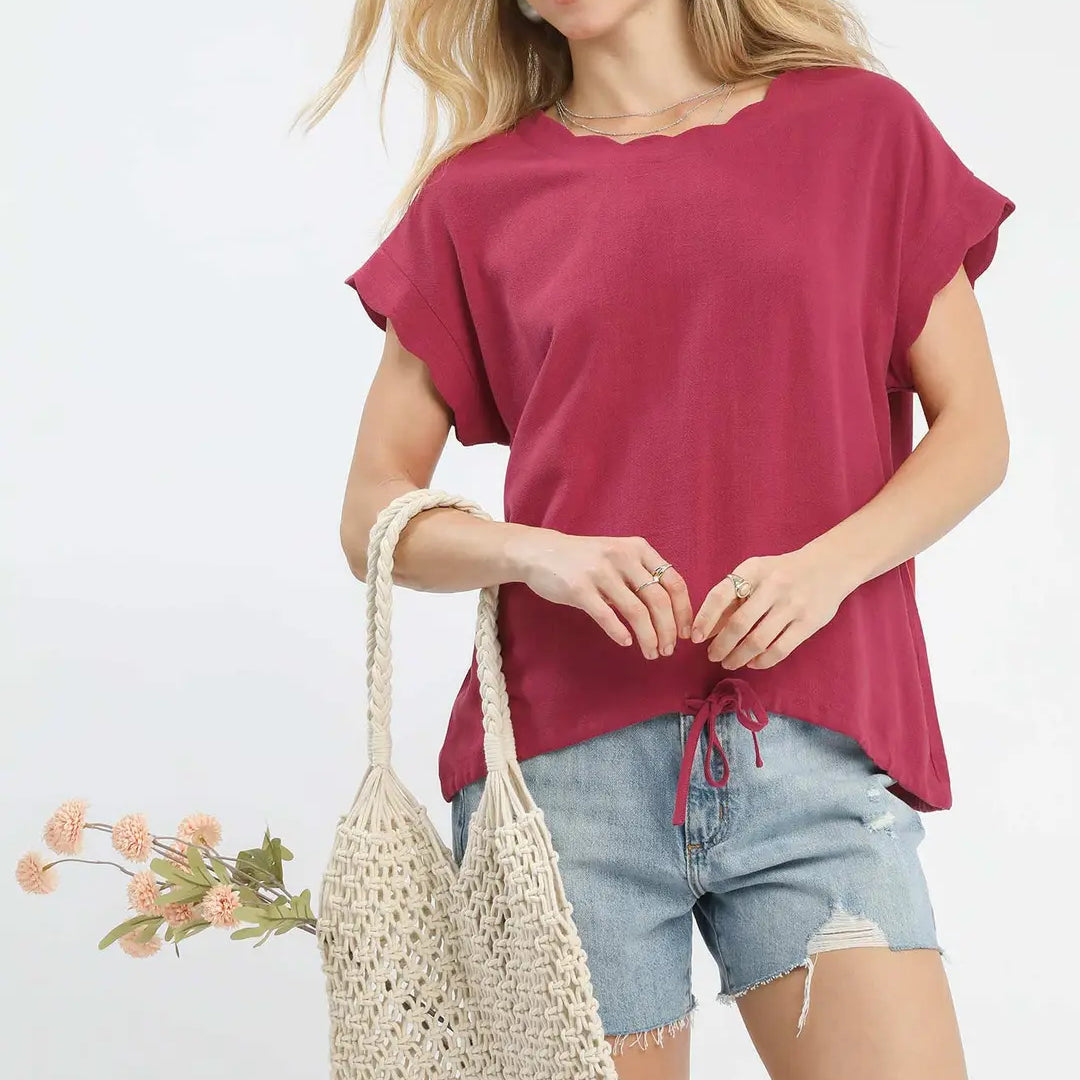 Woman holding a woven bag and wearing a berry-colored top and denim shorts on a white background