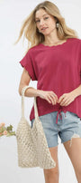 Woman holding a woven bag and wearing a berry-colored top and denim shorts on a white background