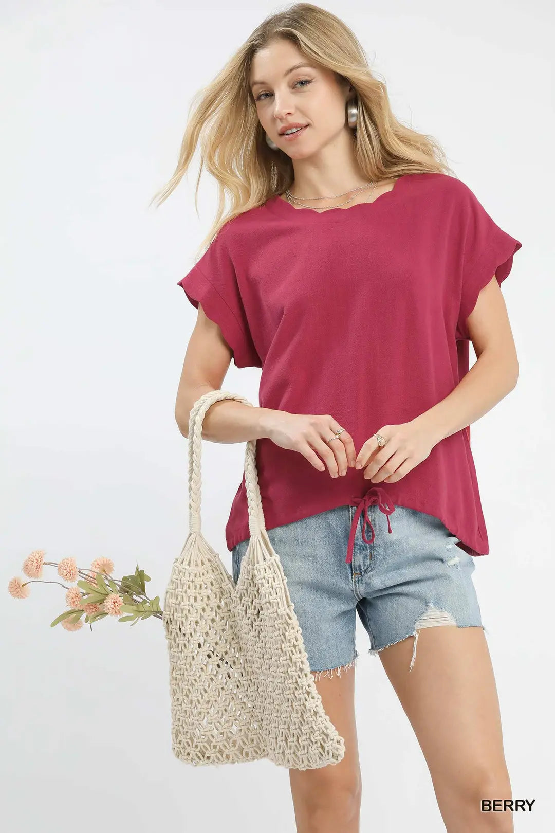 Woman holding a woven bag and wearing a berry-colored top and denim shorts on a white background