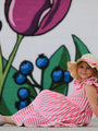 Child in a pink and white striped dress with a straw hat sitting in front of a colorful mural.