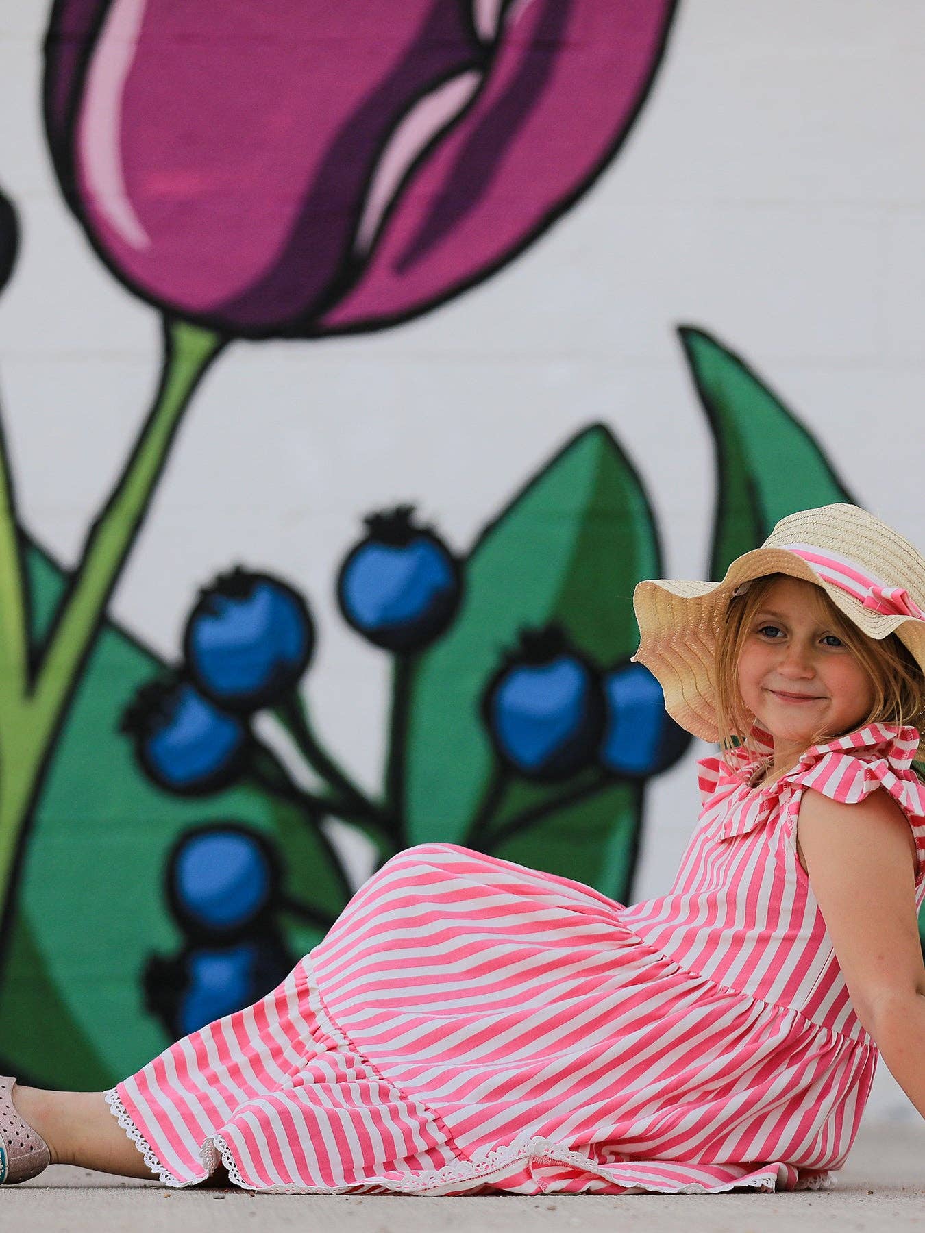 Child in a pink and white striped dress with a straw hat sitting in front of a colorful mural.