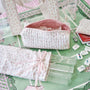 A coordinated set of pastel floral fabric accessories arranged on a green patterned surface, including a quilted zip pouch, a matching bag or scarf-like textile with a pink ribbon, a folded fabric piece, clear plastic tubes, and small white game pieces.