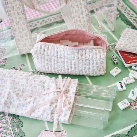 A coordinated set of pastel floral fabric accessories arranged on a green patterned surface, including a quilted zip pouch, a matching bag or scarf-like textile with a pink ribbon, a folded fabric piece, clear plastic tubes, and small white game pieces.