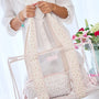 A person in a white blouse holds a pastel floral tote bag with clear plastic front panels and matching small pouches. The bag has a soft pink and cream ditsy floral print, ribbed trim, and a transparent side pocket with a clear strap, set against a bright, softly lit background with flowers on the right.