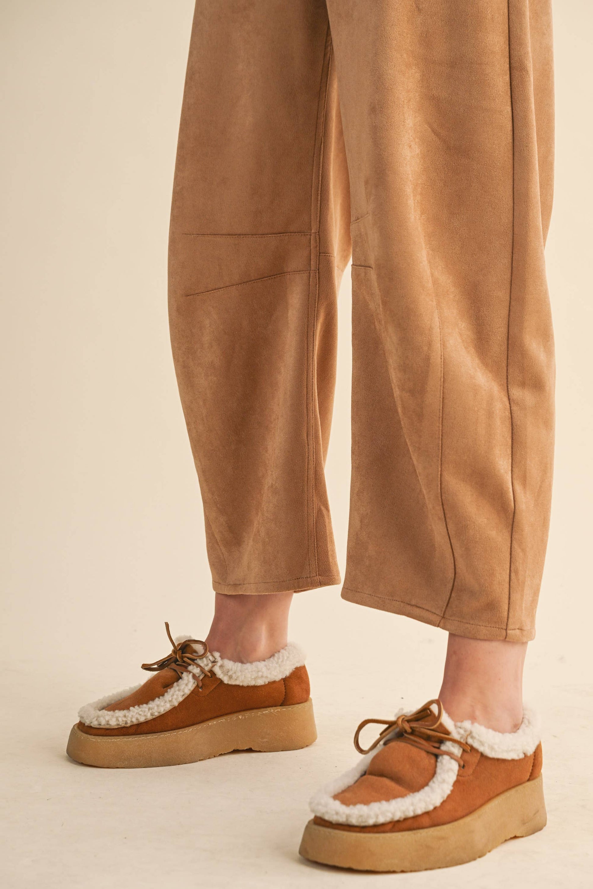 Brown platform shoes with white laces worn with beige pants on a neutral background