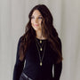 Woman wearing a black long-sleeve top and layered gold necklaces against a neutral background