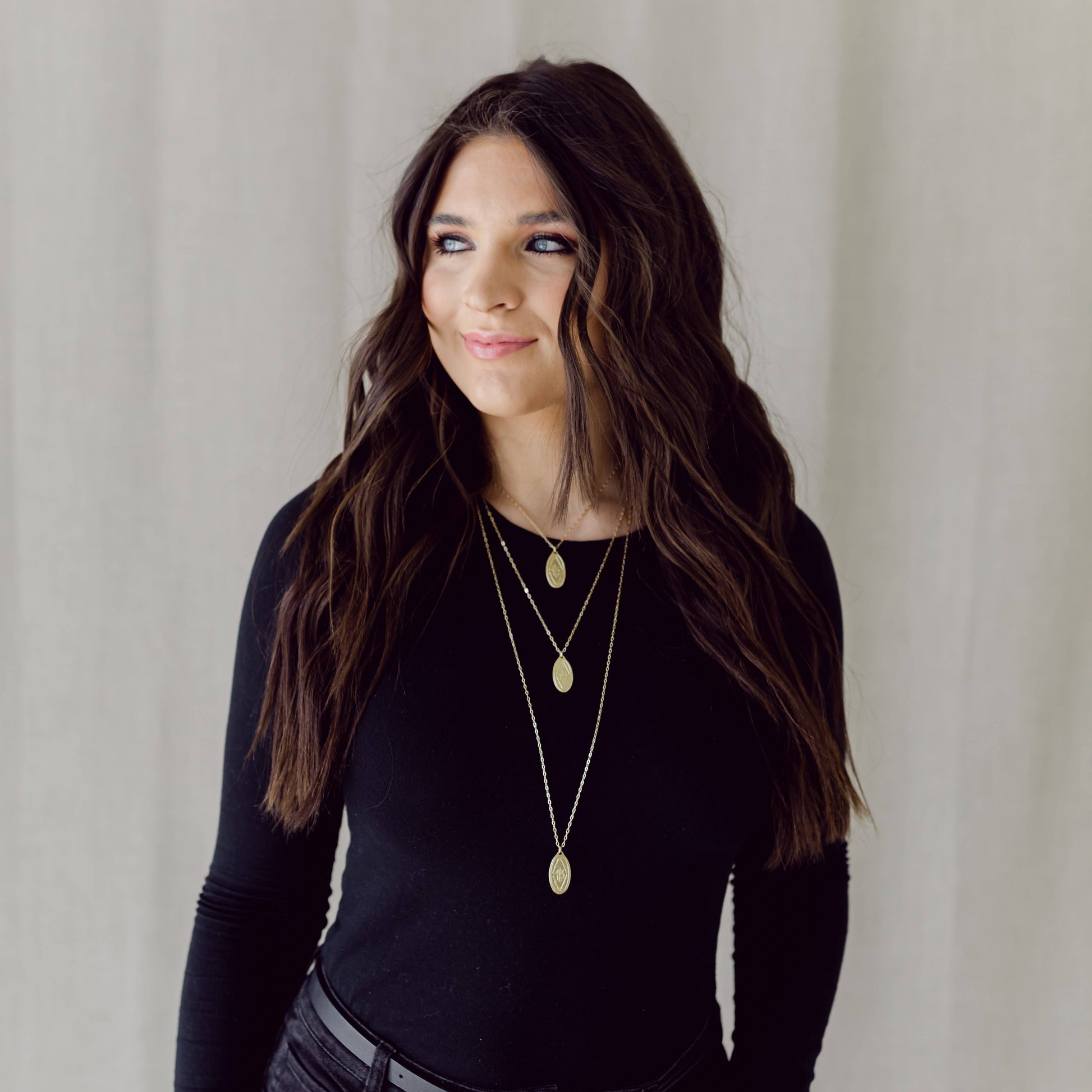Woman wearing a black long-sleeve top and layered gold necklaces against a neutral background