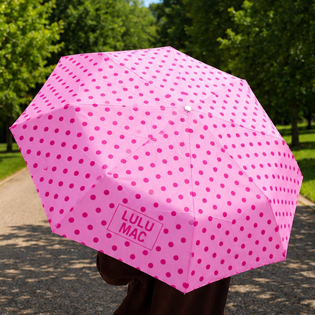 Pink polka dot umbrella with 'LULU MAC' branding held by a person outdoors.