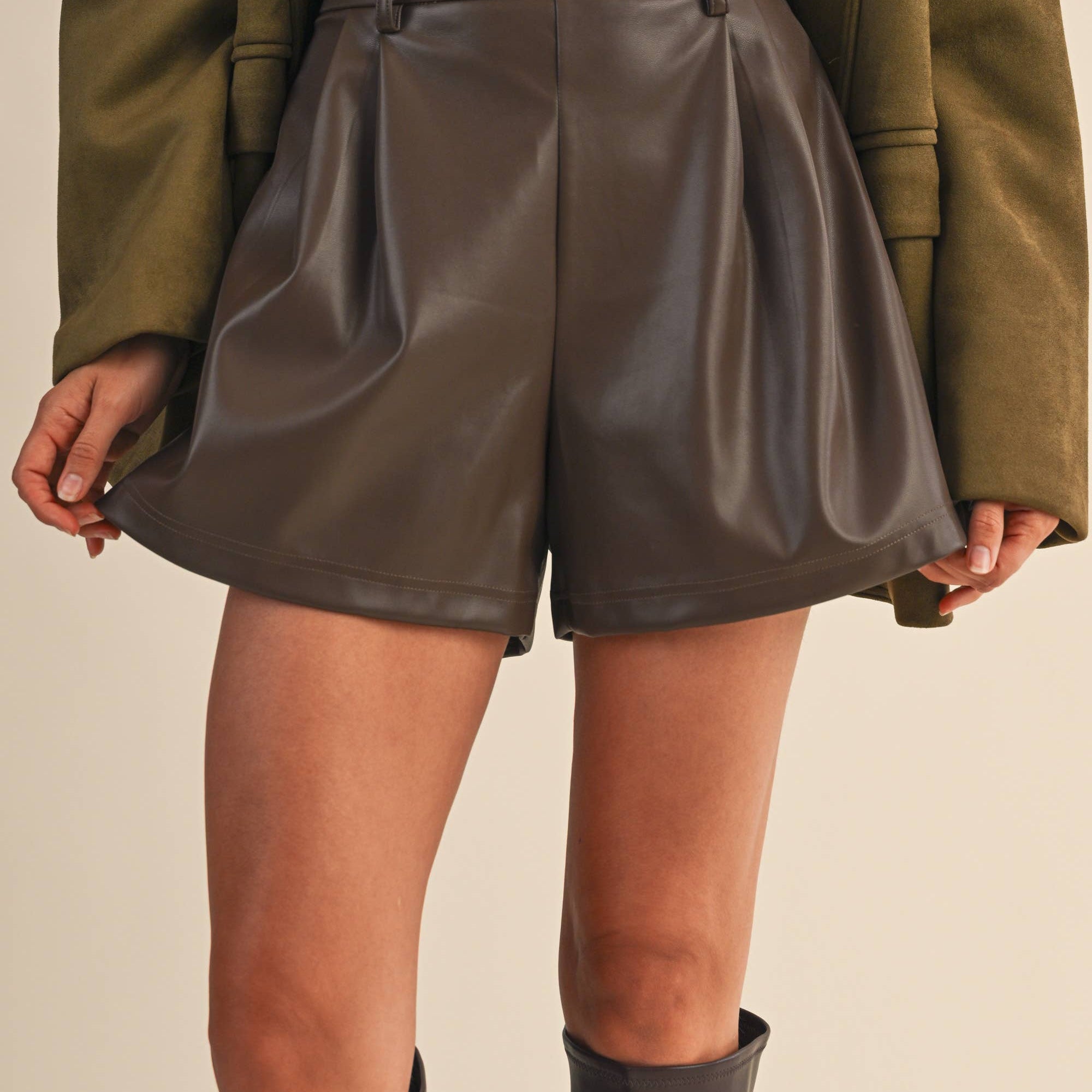 Person wearing brown eather shorts with a green jacket and gray boots on a beige background