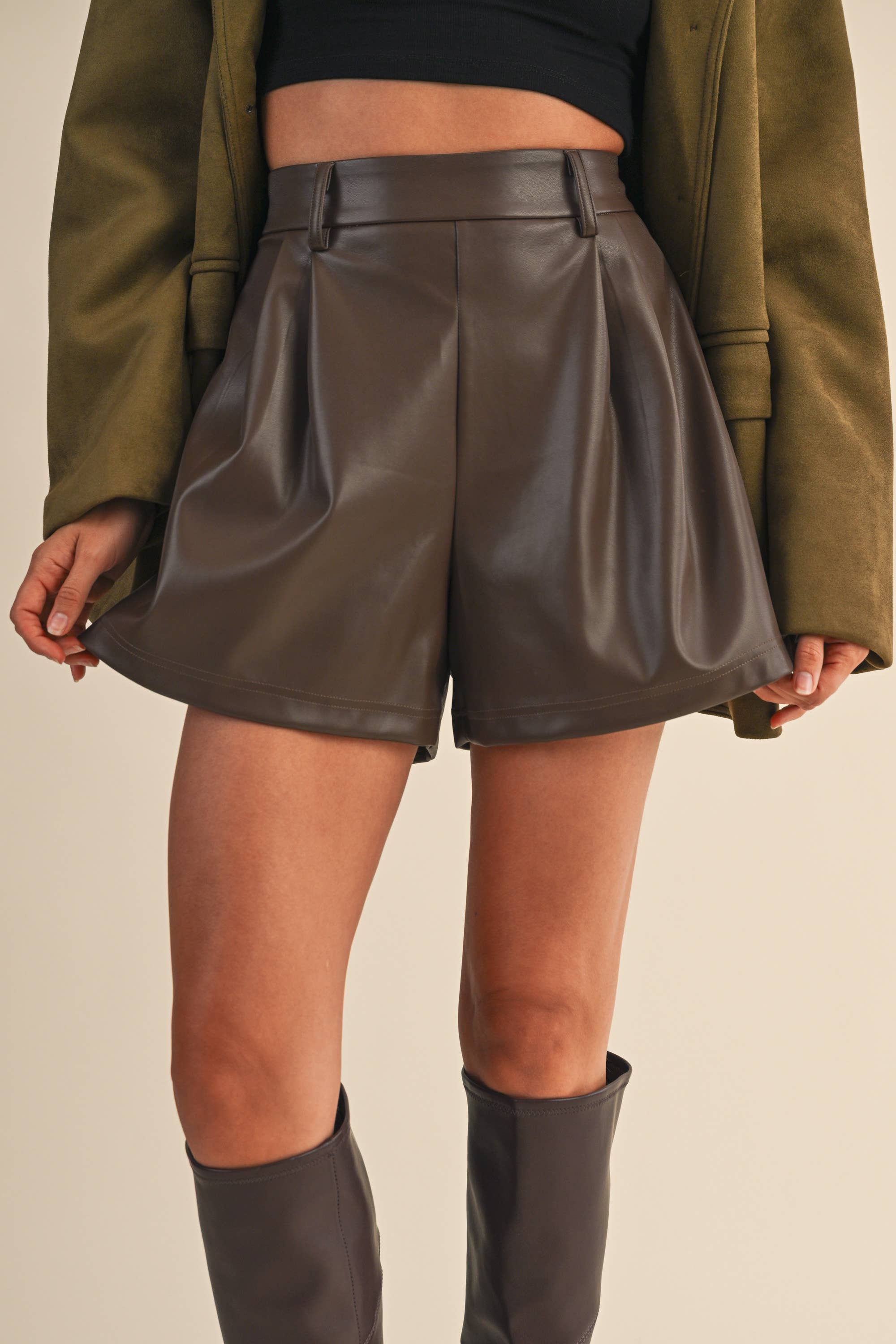 Person wearing brown eather shorts with a green jacket and gray boots on a beige background
