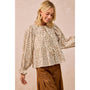 A woman wearing a beige long-sleeve blouse with a small brown floral print, ruffled sleeves, and a drawstring neckline, paired with brown pants, against a soft beige studio background.
