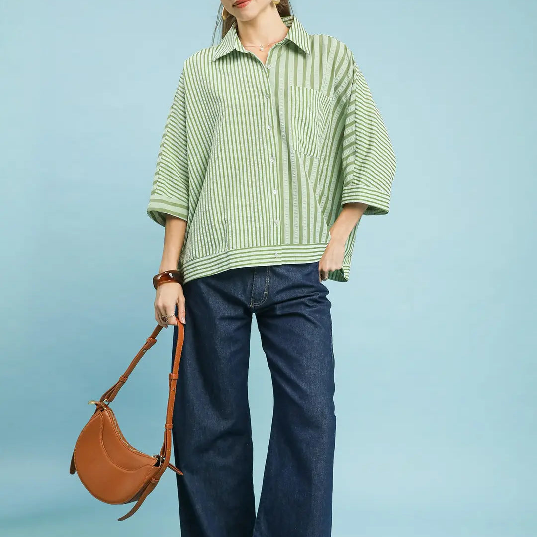 Woman wearing a green striped shirt and blue jeans, holding a brown leather handbag on a light blue background.