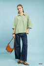 Woman wearing a green striped shirt and blue jeans, holding a brown leather handbag on a light blue background.