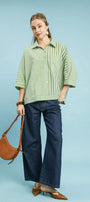 Woman wearing a green striped shirt and blue jeans, holding a brown leather handbag on a light blue background.