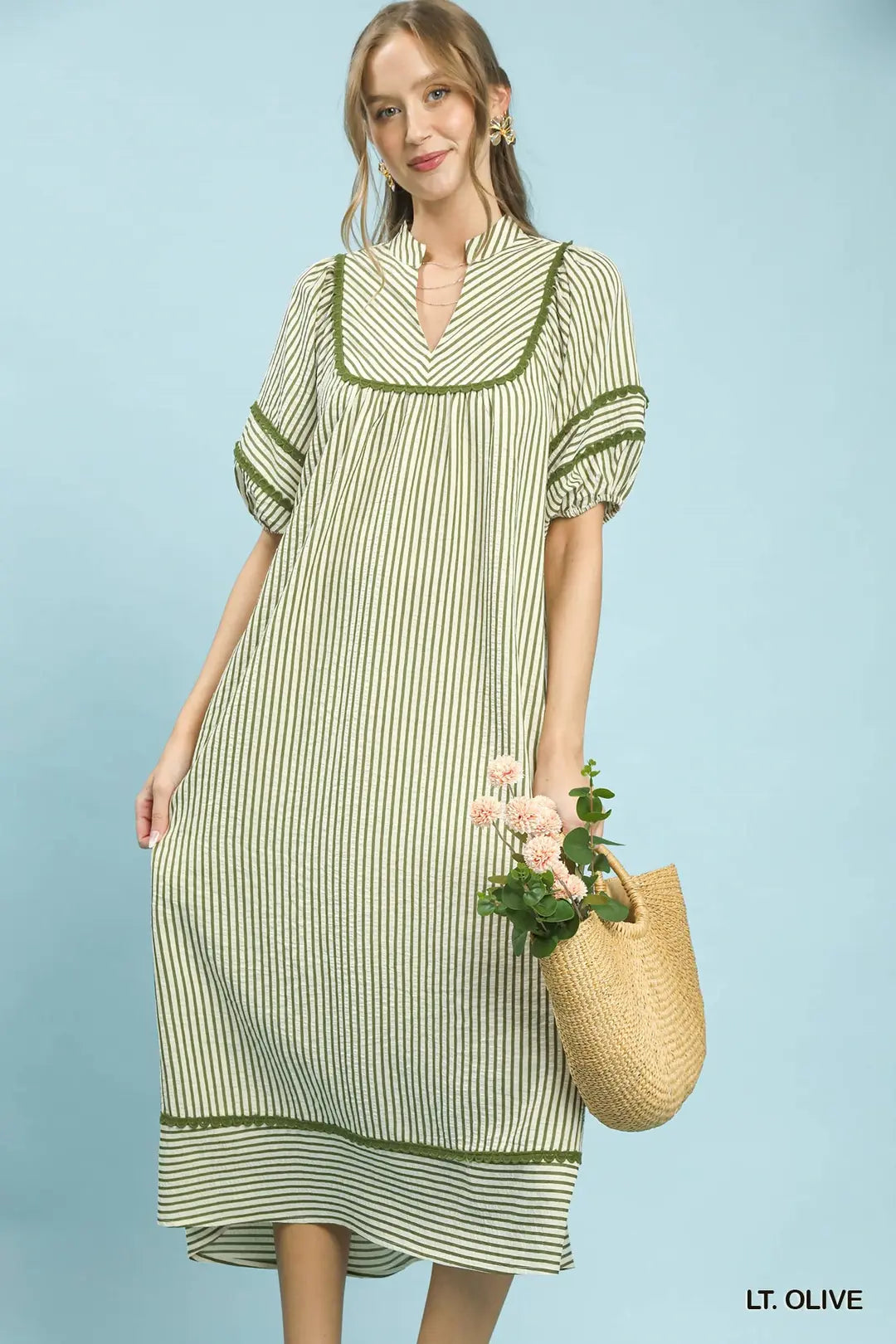 Woman wearing a light olive striped dress holding a straw bag with flowers on a light blue background