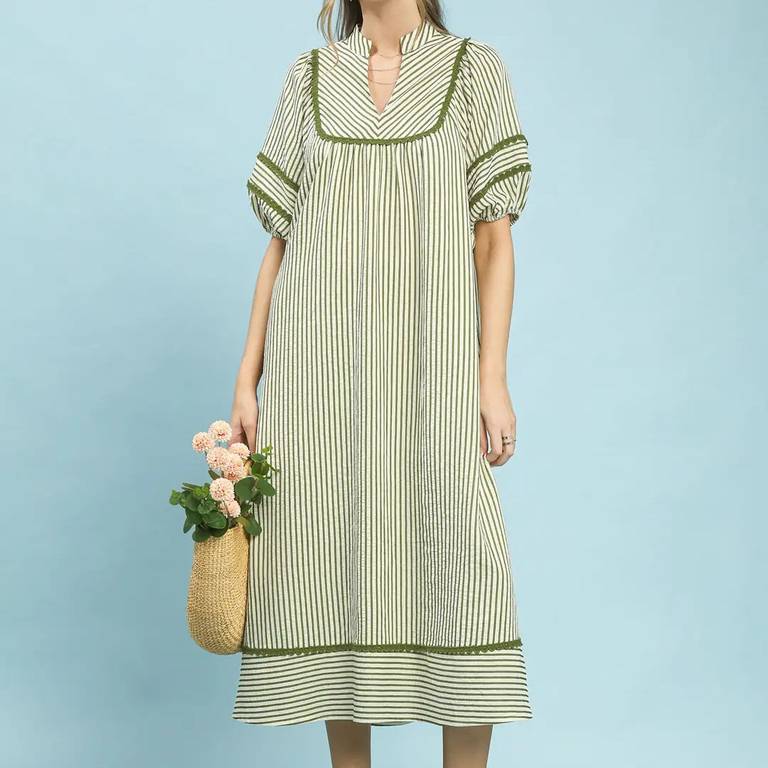 Woman wearing a light olive striped dress holding a woven bag against a light blue background