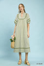 Woman wearing a light olive striped dress holding a woven bag against a light blue background