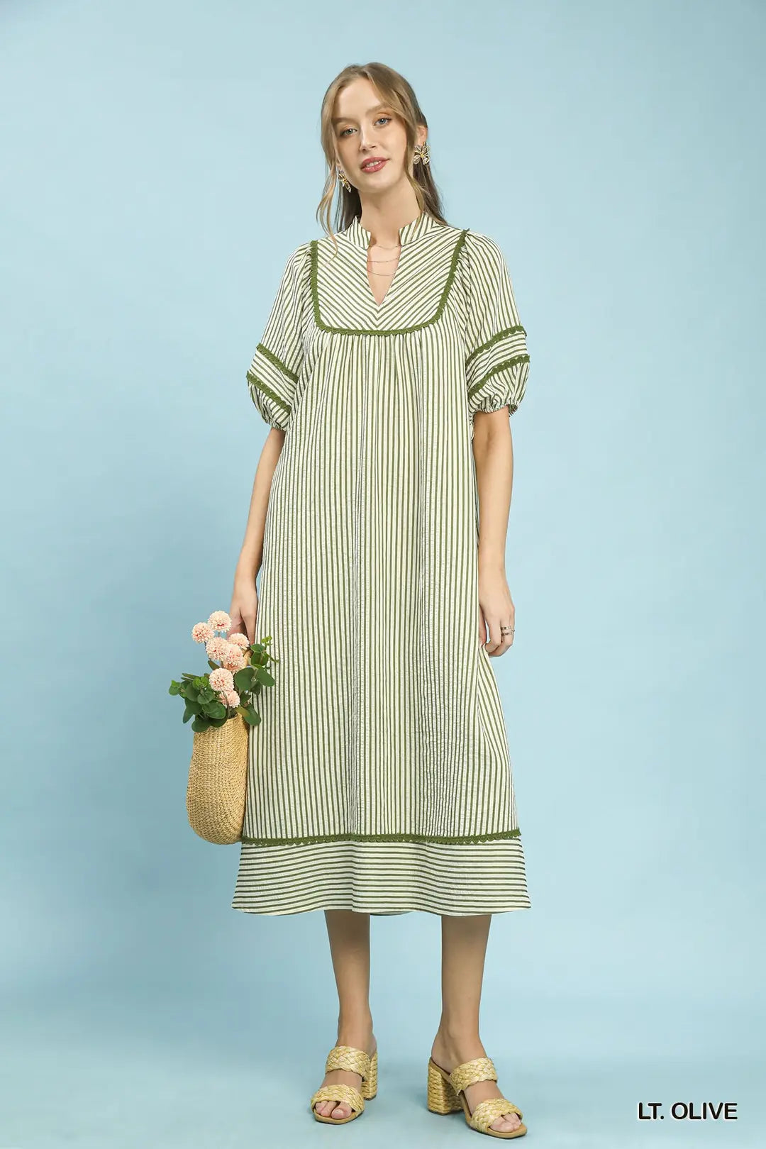Woman wearing a light olive striped dress holding a woven bag against a light blue background