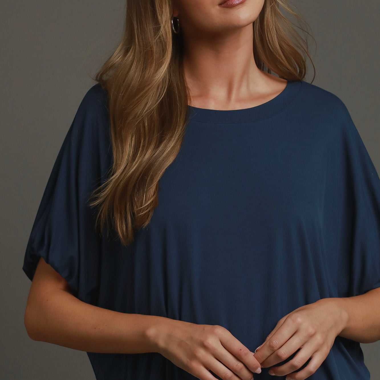 A woman wearing a navy blue short-sleeve top with a relaxed, draped fit and white pants, posed against a solid dark gray studio background.