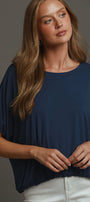 A woman wearing a navy blue short-sleeve top with a relaxed, draped fit and white pants, posed against a solid dark gray studio background.