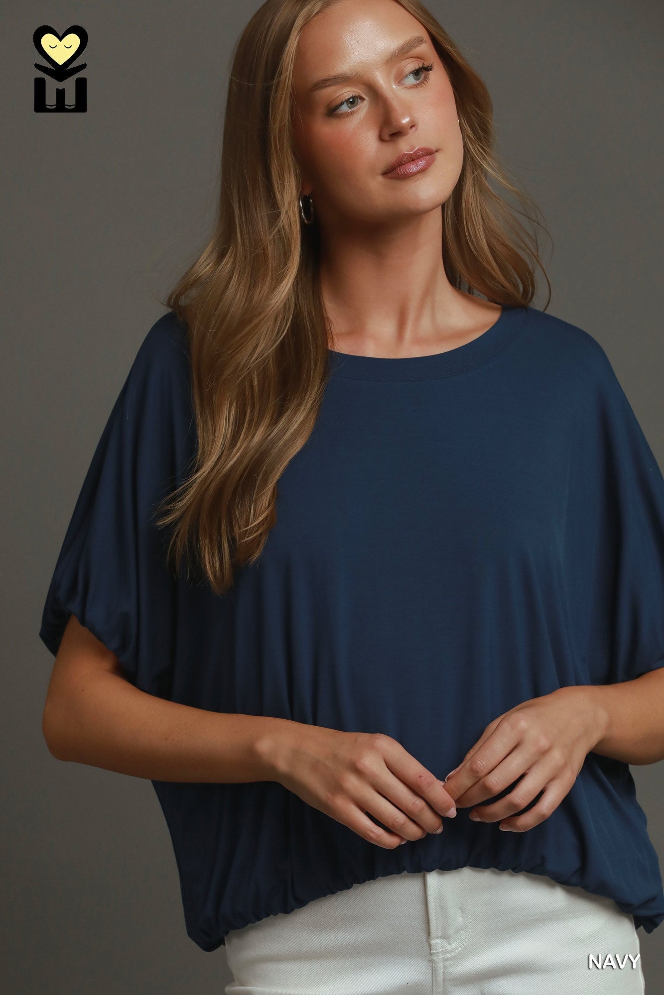 A woman wearing a navy blue short-sleeve top with a relaxed, draped fit and white pants, posed against a solid dark gray studio background.