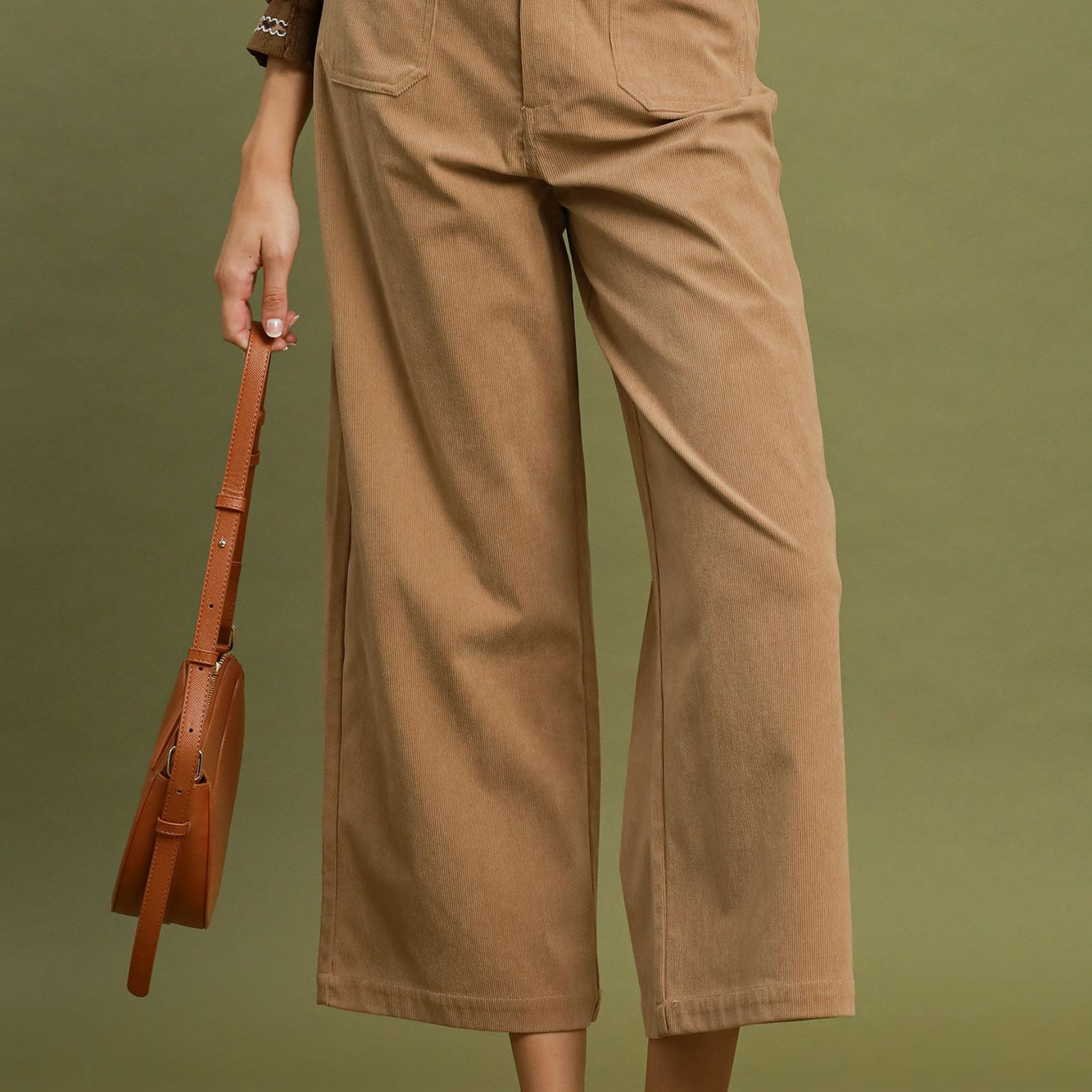A model wearing taupe wide-leg cropped pants with a matching brown belt, paired with a brown top and brown pointed-toe mule heels, holding a brown leather shoulder bag against an olive green background.