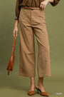 A model wearing taupe wide-leg cropped pants with a matching brown belt, paired with a brown top and brown pointed-toe mule heels, holding a brown leather shoulder bag against an olive green background.