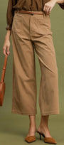 A model wearing taupe wide-leg cropped pants with a matching brown belt, paired with a brown top and brown pointed-toe mule heels, holding a brown leather shoulder bag against an olive green background.