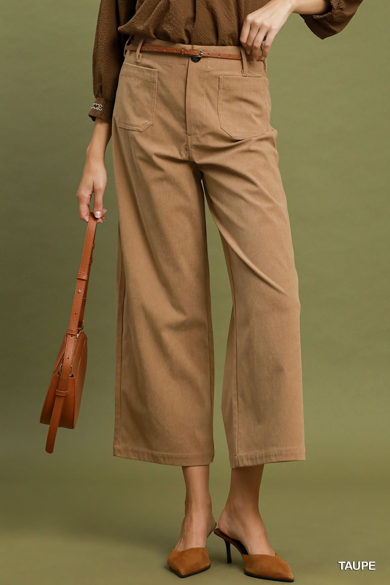 A model wearing taupe wide-leg cropped pants with a matching brown belt, paired with a brown top and brown pointed-toe mule heels, holding a brown leather shoulder bag against an olive green background.