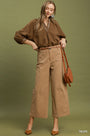 A woman modeling a brown long-sleeve button-up shirt and taupe wide-leg cropped pants, styled with a brown belt, brown pointed-toe heels, and a brown shoulder bag against an olive green studio background.