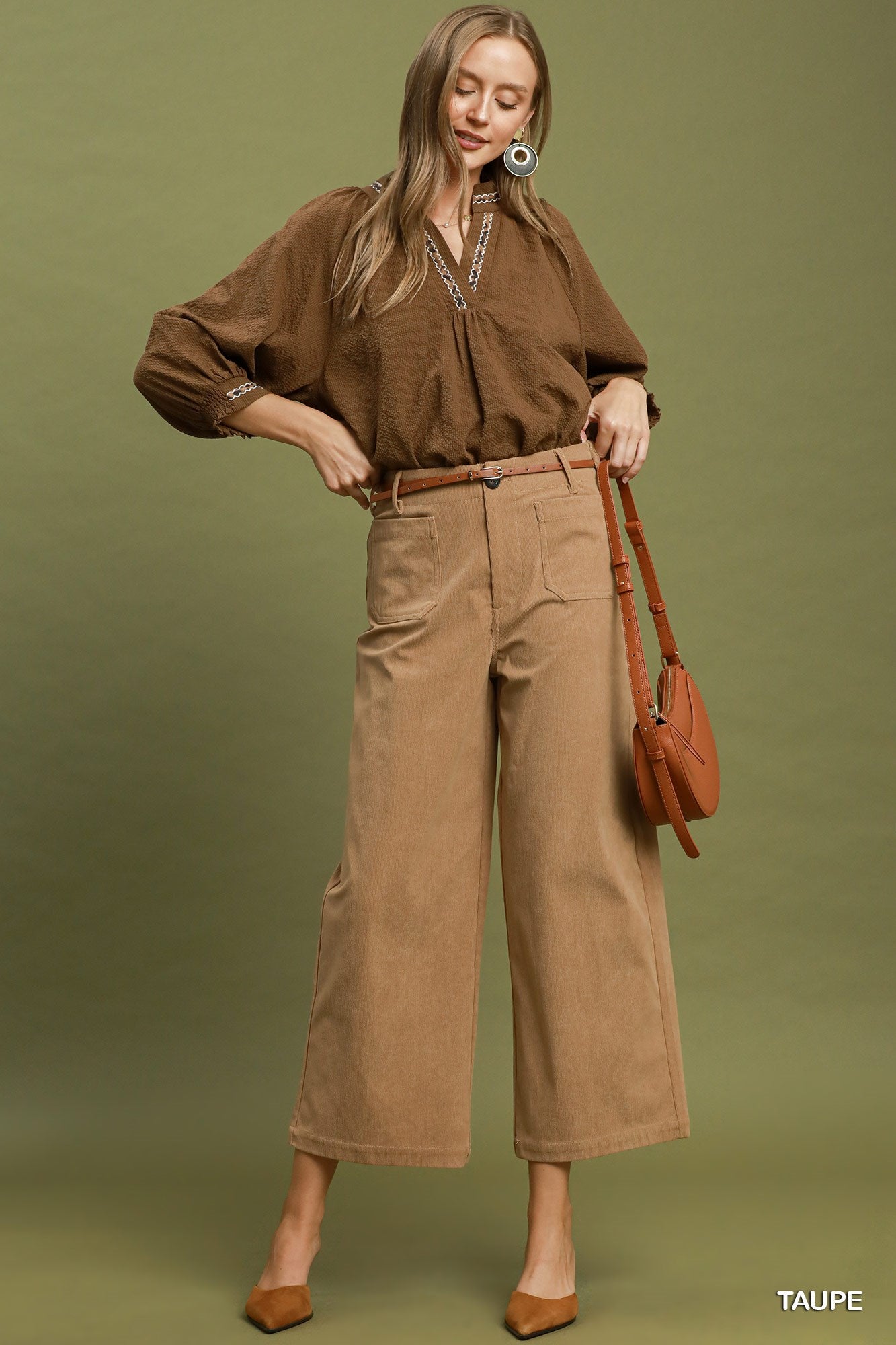 A woman modeling a brown long-sleeve button-up shirt and taupe wide-leg cropped pants, styled with a brown belt, brown pointed-toe heels, and a brown shoulder bag against an olive green studio background.