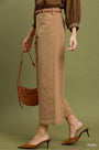 A model wearing taupe wide-leg cropped trousers, a brown long-sleeve blouse, and brown mule heels, holding a matching brown crescent handbag against a solid olive green background.