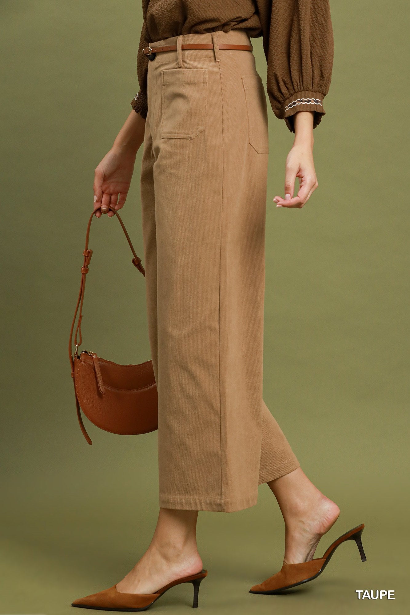 A model wearing taupe wide-leg cropped trousers, a brown long-sleeve blouse, and brown mule heels, holding a matching brown crescent handbag against a solid olive green background.