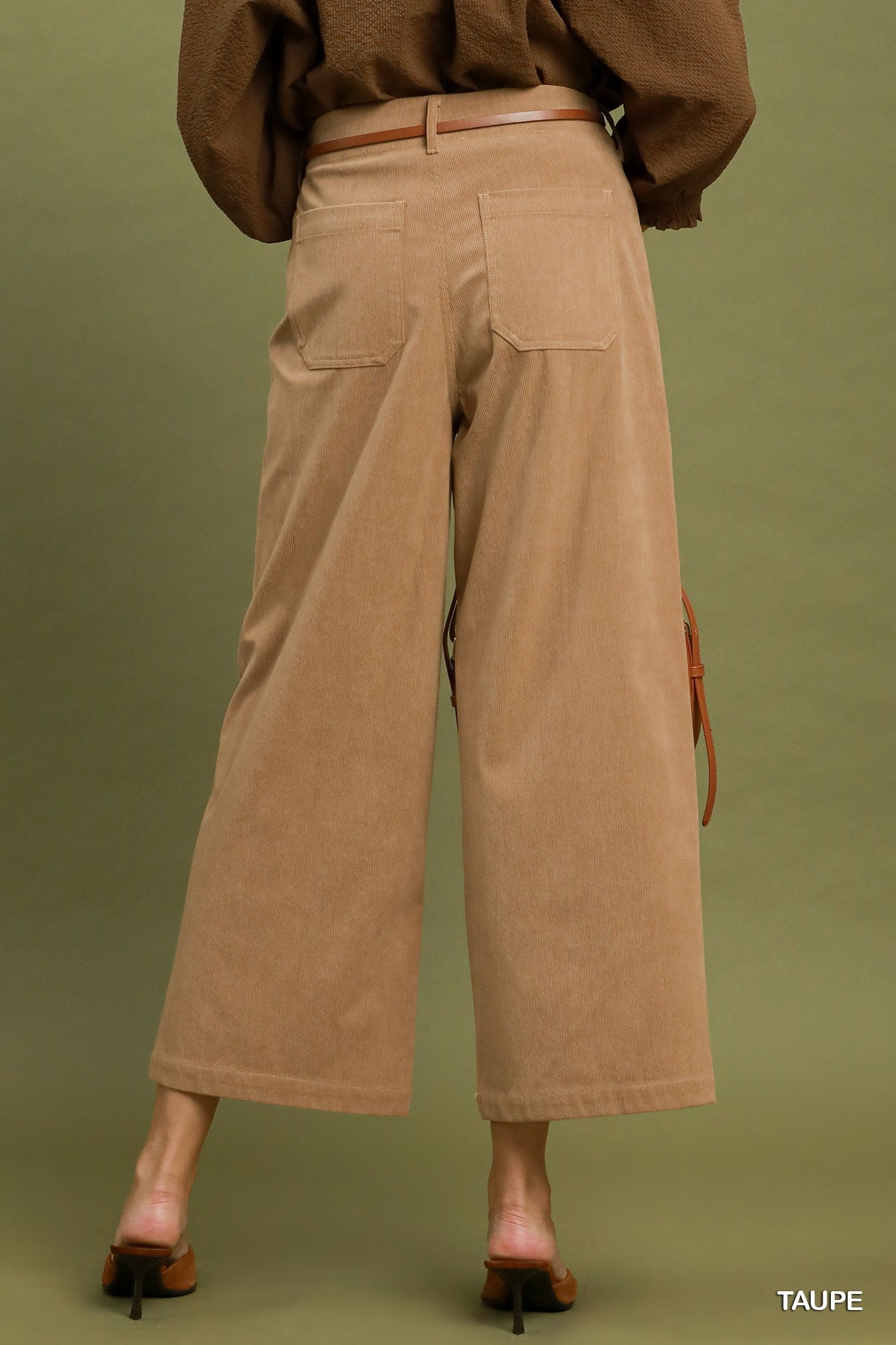 A person shown from the back wearing wide-leg cropped pants in taupe, paired with a brown belt and brown heeled mule sandals, against an olive green studio background.