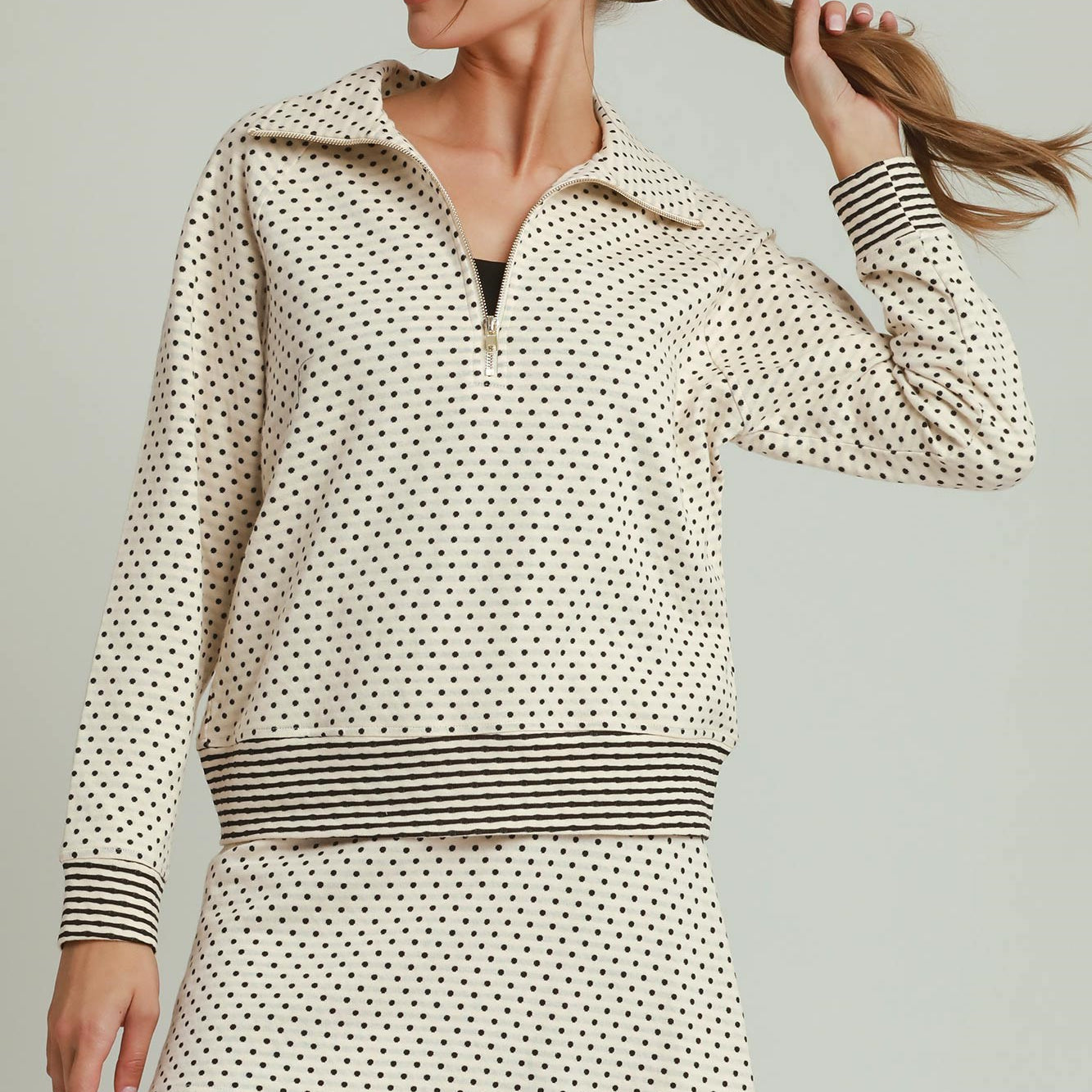 A woman modeling a cream and black mini dress with a small polka dot pattern, a half-zip collar, and striped cuffs and waistband, set against a solid light gray background.