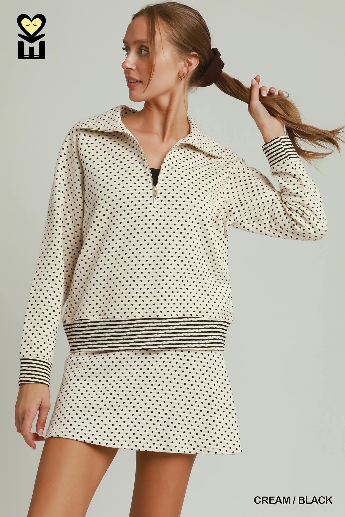 A woman modeling a cream and black mini dress with a small polka dot pattern, a half-zip collar, and striped cuffs and waistband, set against a solid light gray background.