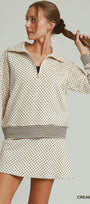 A woman modeling a cream and black mini dress with a small polka dot pattern, a half-zip collar, and striped cuffs and waistband, set against a solid light gray background.