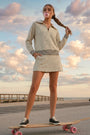 A woman stands on a skateboard outdoors near a boardwalk and ocean under a cloudy sky, wearing a beige and black polka-dot long-sleeve mini dress with a striped waist detail and black sneakers.