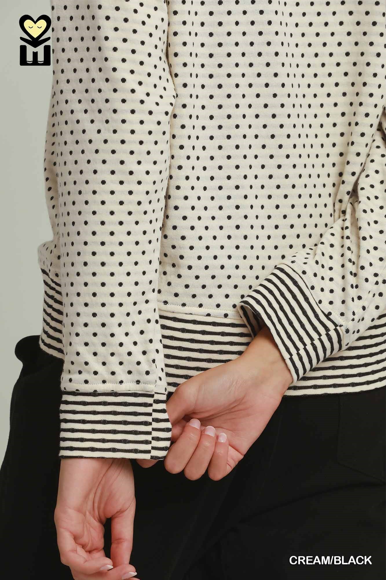 Close-up of a cream-colored long-sleeve top with black polka dots and black-and-white striped cuffs and hem, worn by a model.