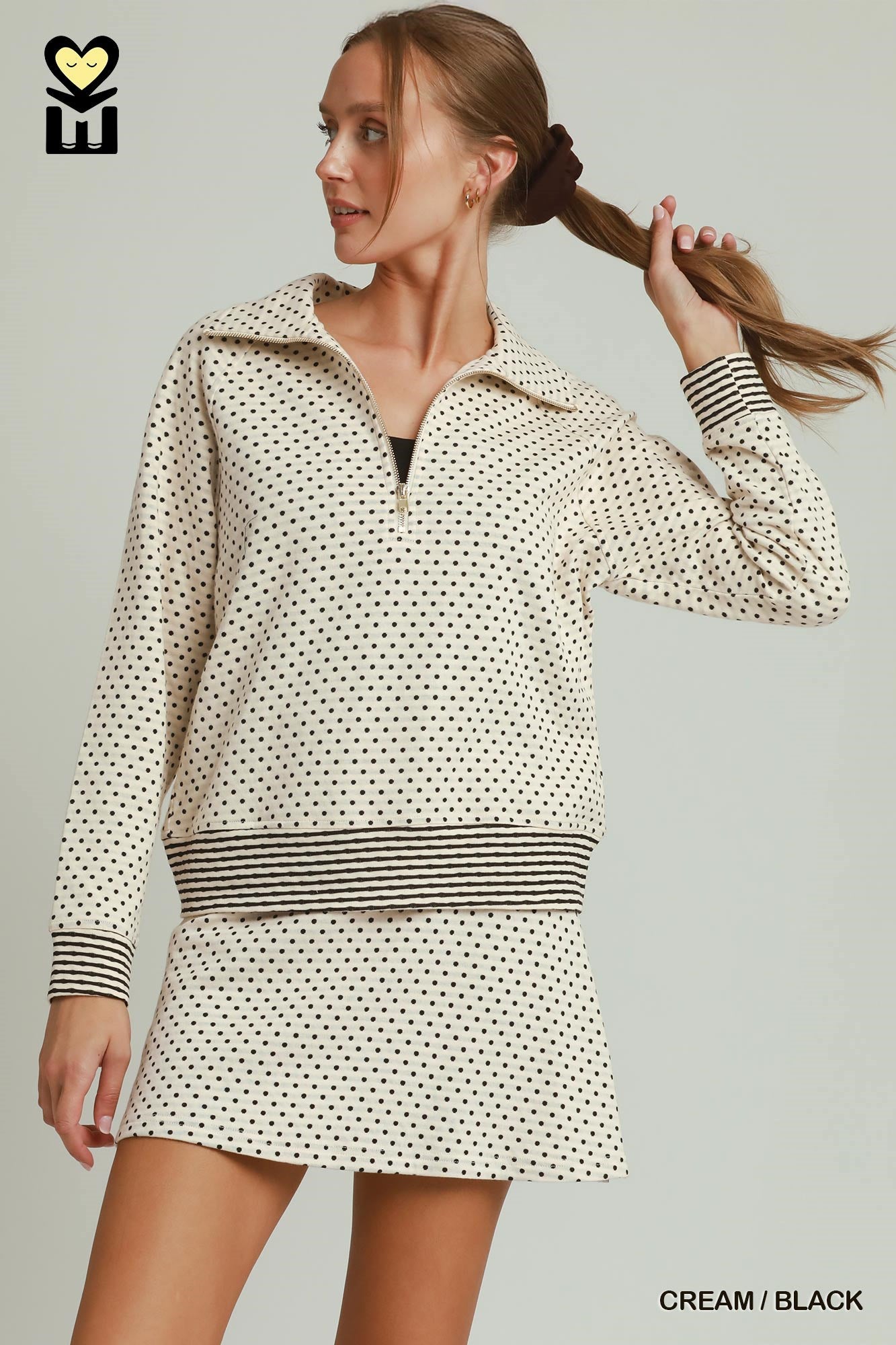 A woman modeling a cream and black mini dress with a small polka dot pattern, a half-zip collar, and striped cuffs and waistband, set against a solid light gray background.